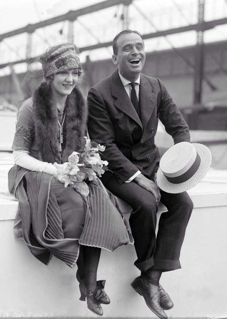 the big band era's influence on fashion - couple in the 1930's sitting on a concrete fence dressed in elegant day wear and laughing