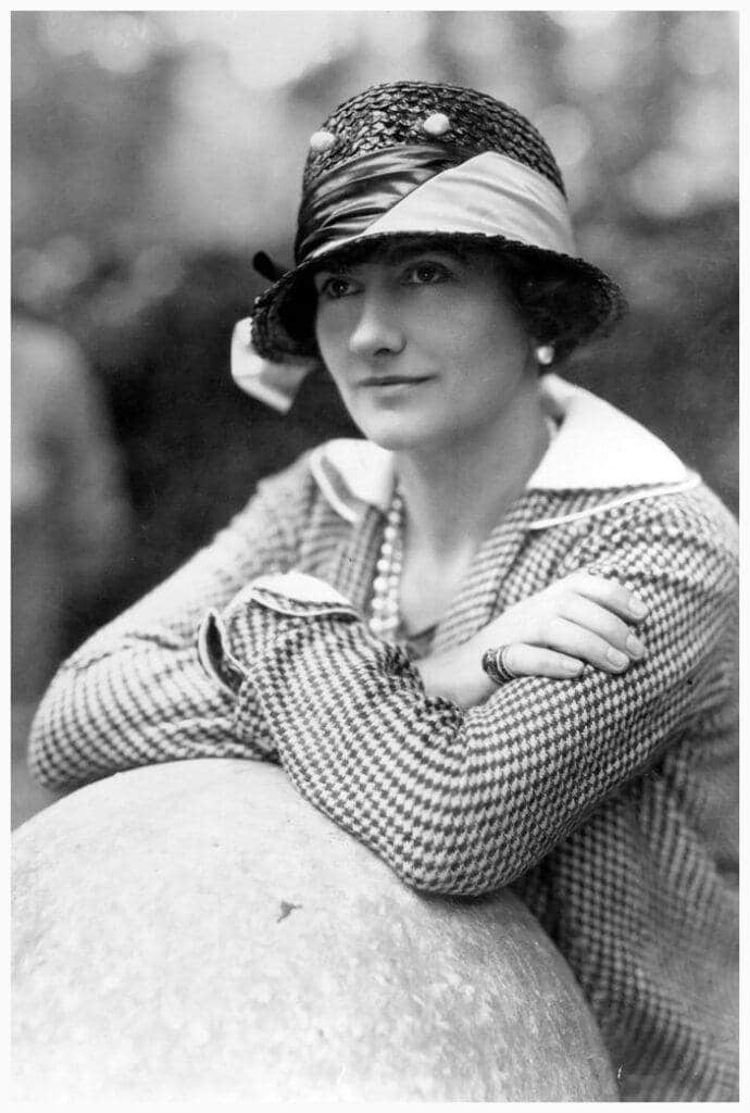 the impact of fashion's golden age icons - coco chanel leaning on a statue in a 1930's hat