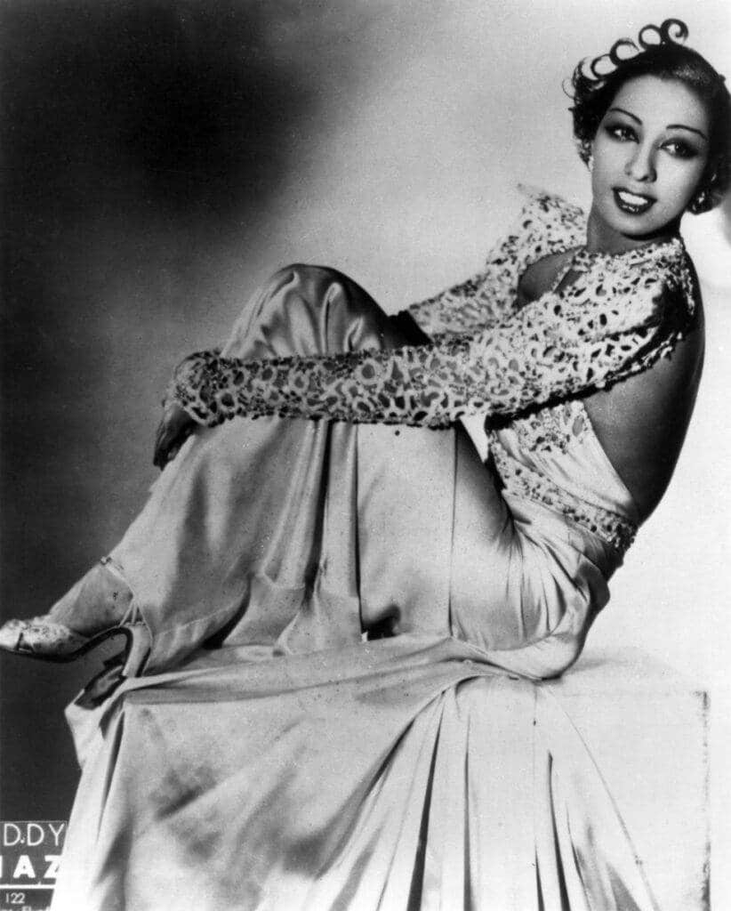 the impact of fashion's golden age icons - josephine baker perched on a table draped in satin dress with animal print sleeves