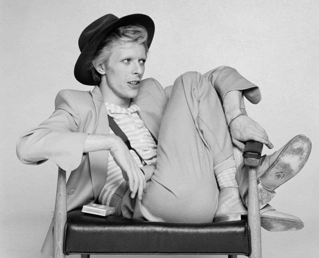 the impact of fashion's golden age icons - david bowie huddled onto an arm chair wearing a loose suit and hat