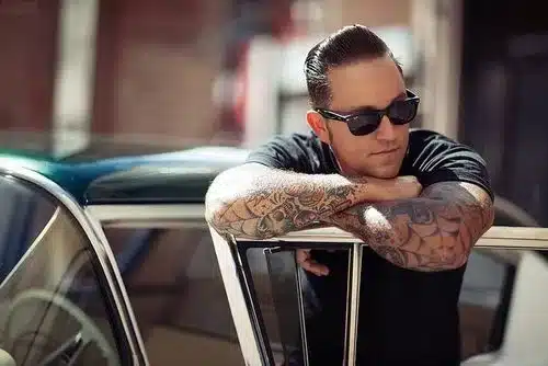 reviewing the essence of rockabilly style for men - man leaning over vintage car with sunnies, tatts and black glasses
