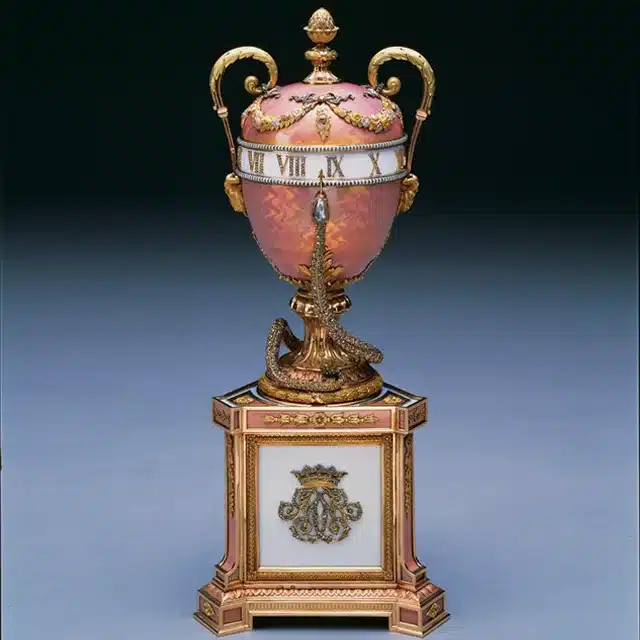 introduction to the allure of faberge eggs - the duchess of marlborough egg circa 1902 egg on top of a podium in pink shades with gold embossing and roman numerals around the top