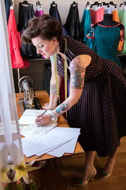 styling tips for pulling off a rockabilly look with modern flair at the shops - woman designing a costume on paper sporting tattoos and fitted dress