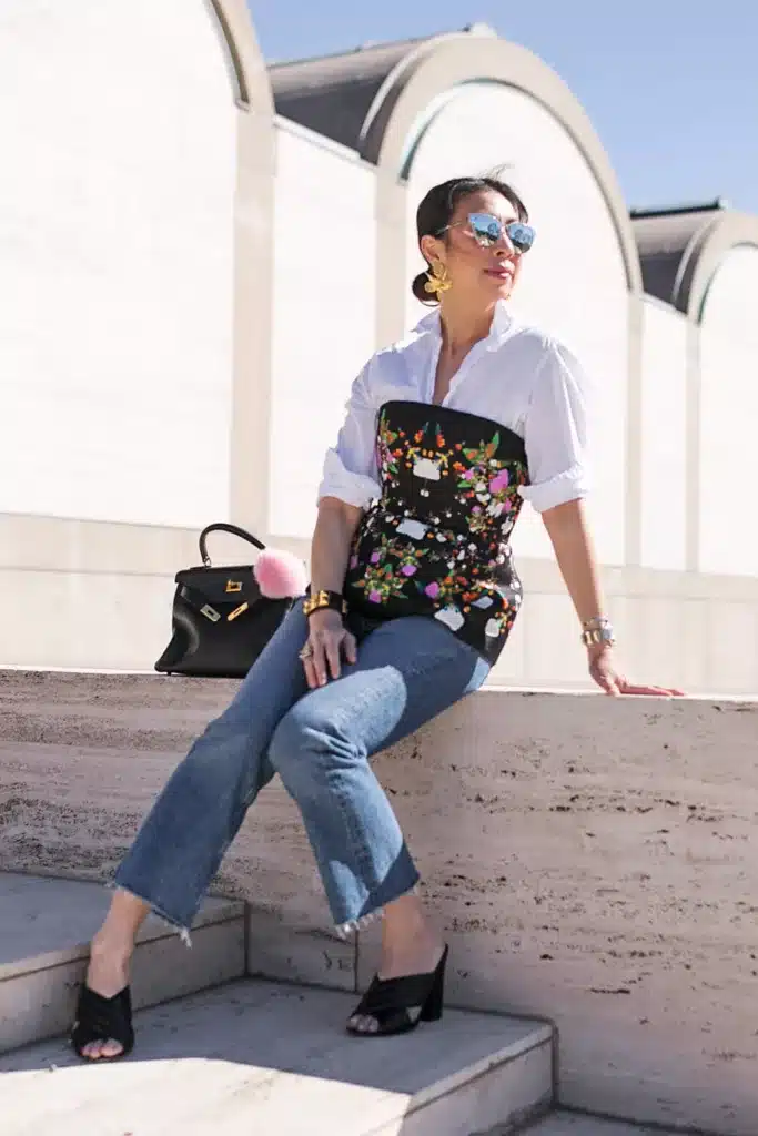 how to incorporate corsets into modern wardrobes - woman sitting on a stone pillar wearing an outside floral corset jeans and white shirt