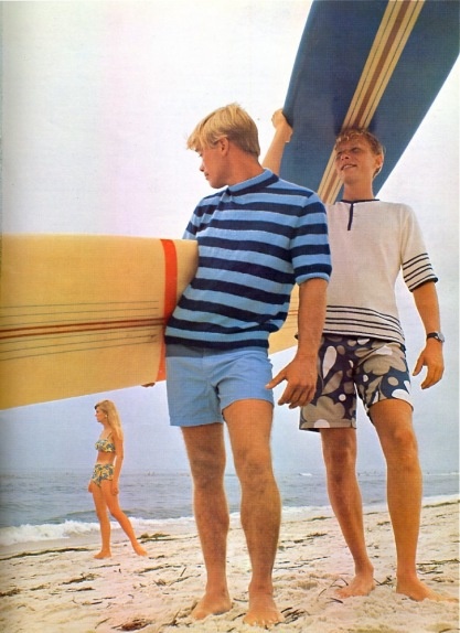 2 blonde surfers on the beach carrying boards wearing longer trunks and striped t.shirts