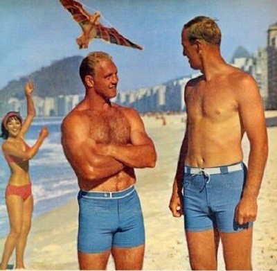 2 blonde guys talking on the beach watching a bird fly high in blue fitted swimming trunks