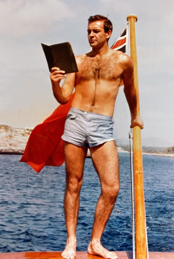 Sean Connery in brief swimming trunks reading a book holding a british flag on a beach