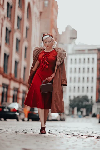 woman walking on city street with a red dress and over coat with hat fascinator gloves and closed shoes