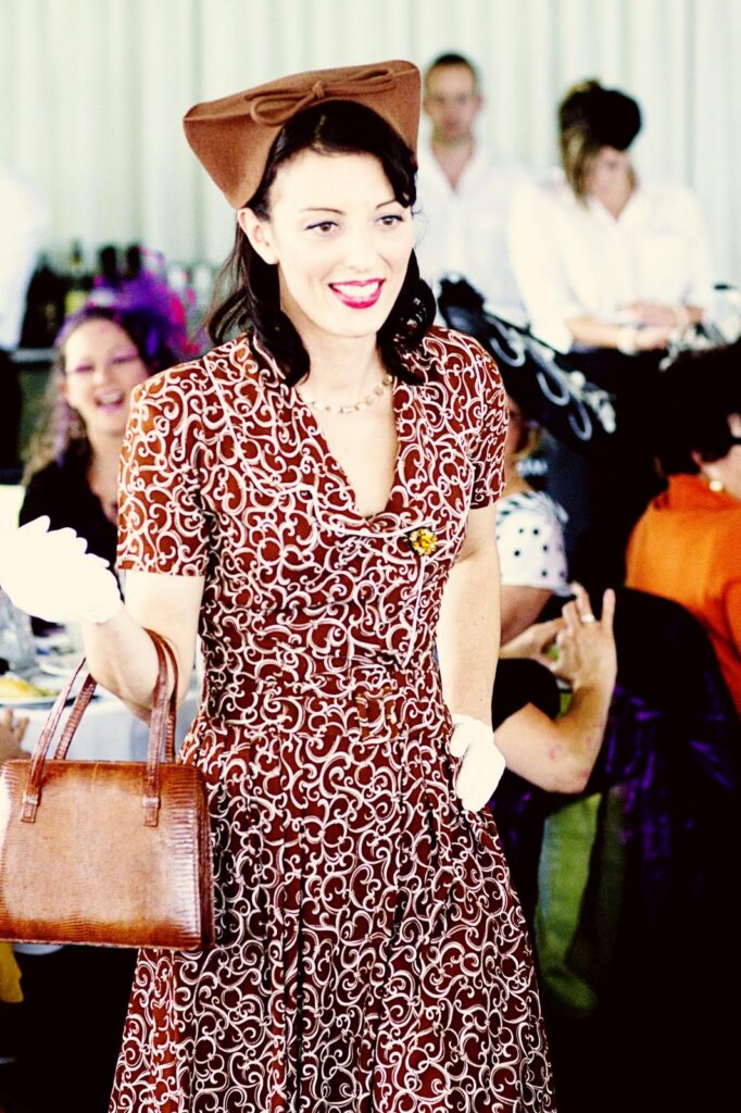 Lady dressed in 1940's patterned dress with brown handbag gloves and U shape pill box hat