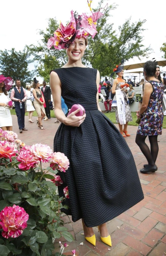 Lady dressed in black boat neck sleeveless dress pleated at the waist with big floral fascinator and contrast yellow court shoes