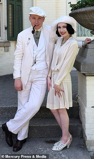 Couple dressed in white and off white outfits man in 3 piece suit with club shirt and newsboy hat and pipe and lady in 1920's loose dress with sunhat to match