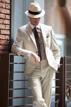 Man posing in a off white suit with striped shirt, trilby hat and suit accessories