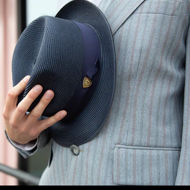 Man holding a navy fedoran straw hat against a grey striped suit