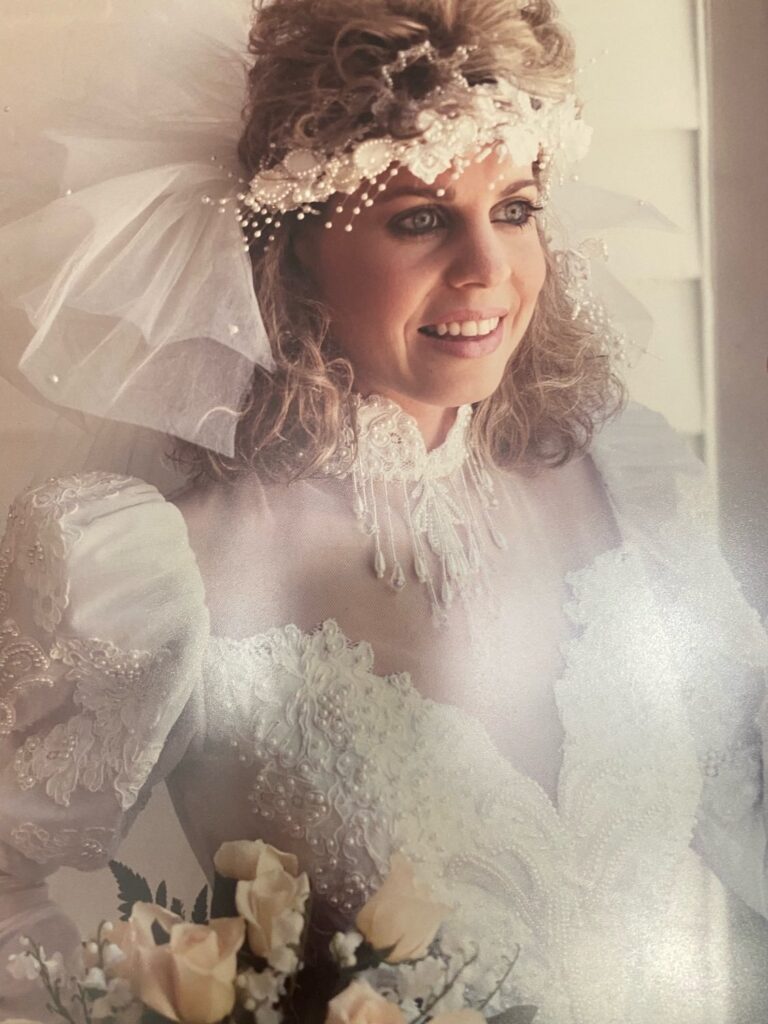 Bride holding beige roses dressed in a long sleeved bead encrusted dress with sweetheart mesh panel and beaded headband with tulle poking out around teased high hair do