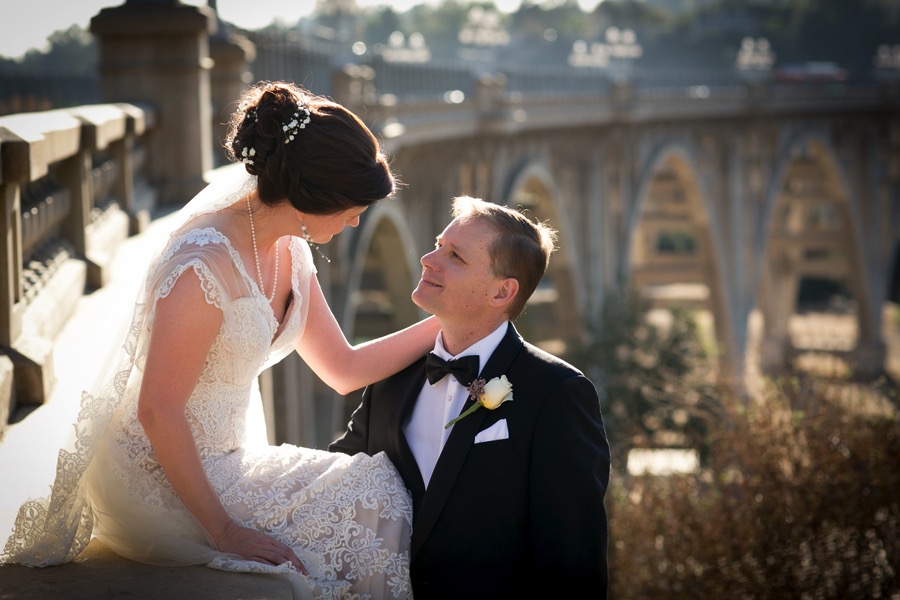 Couple on a bridge in vintage inspired tuxedo attire and lace wedding dress with updo and veil trailing behind