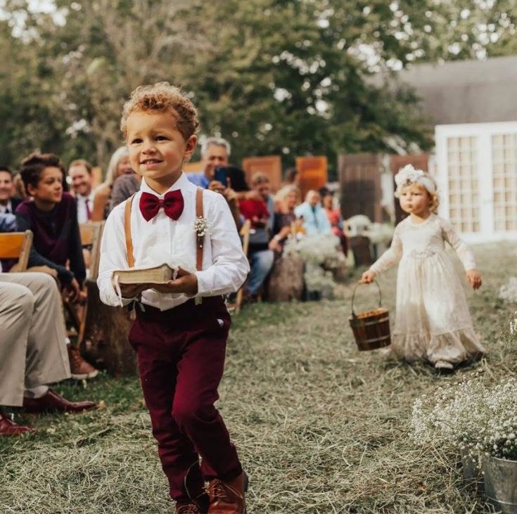 Little boy walking in front of a flower girl holding a book dressed in brown suspenders and burgundy pants and bow tie