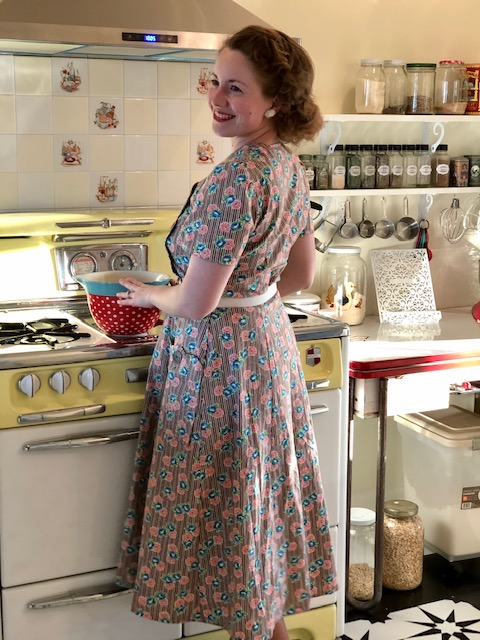 lady standing in the kitchen holding a bowl wearing a blue and pink 1950's house dress