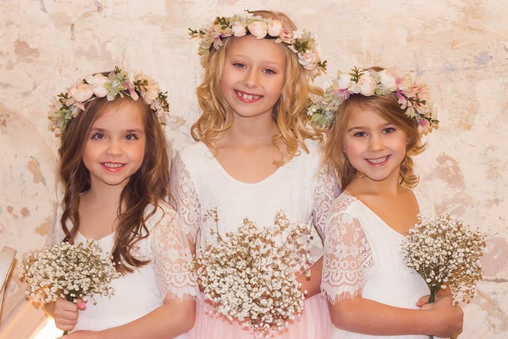 3 girls in identical white dresses with lace sleeves wearing thick flower hair garlands and holding gip flowers