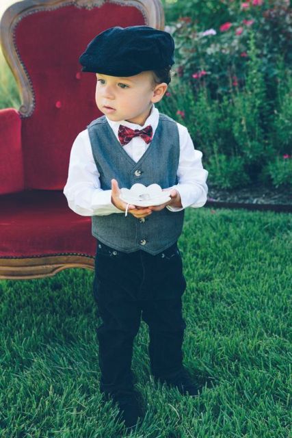 Little boy holding a ring tray wearing grey vest and white shirt with a tartan bow tie and black newsboy cap