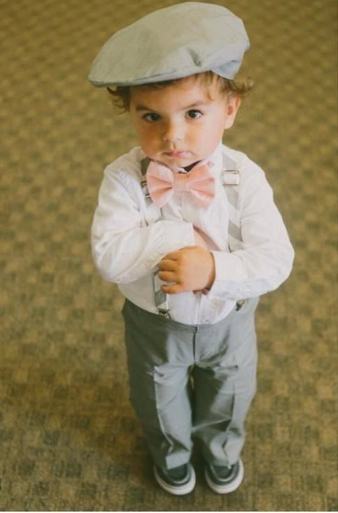 Little boy in grey pants and silver suspenders with a pink bow tie and newsboy cap holding something in his hands