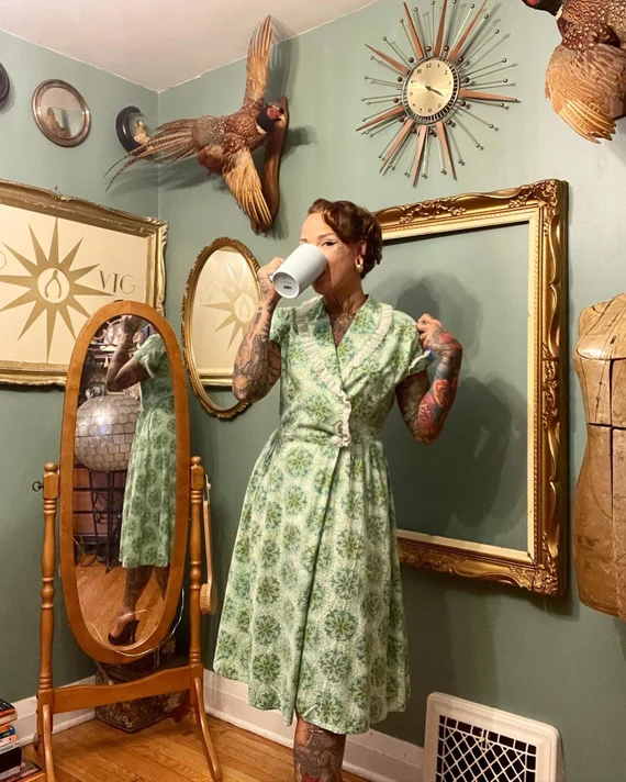 lady drinking a cup of tea wearing a green and beige house dress in her living room adorned with wall decor