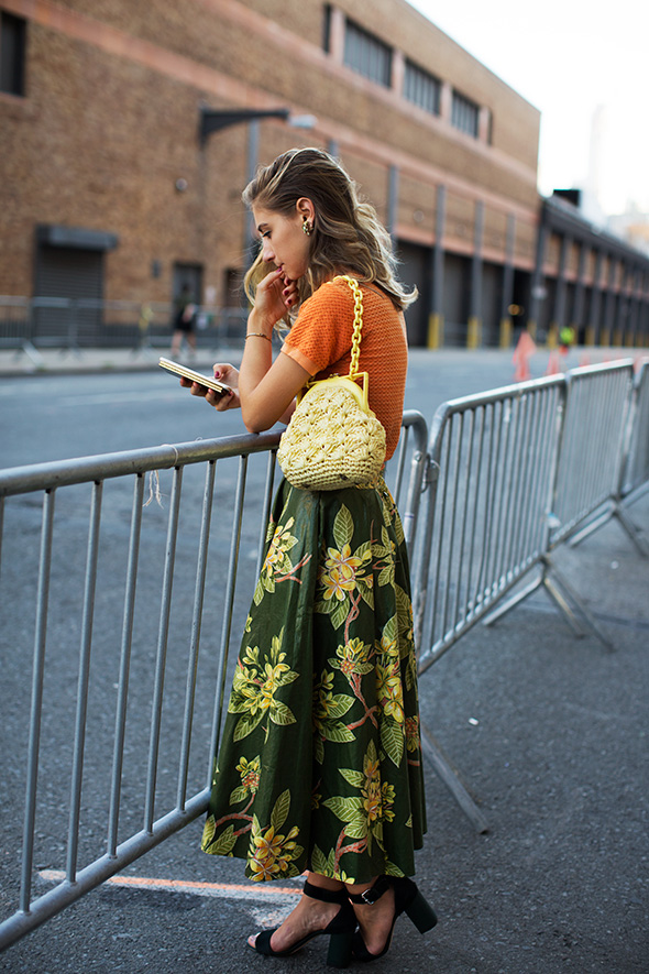 woman standing on a street wearing a green and gold vintage skirt and orange top with lace handbag