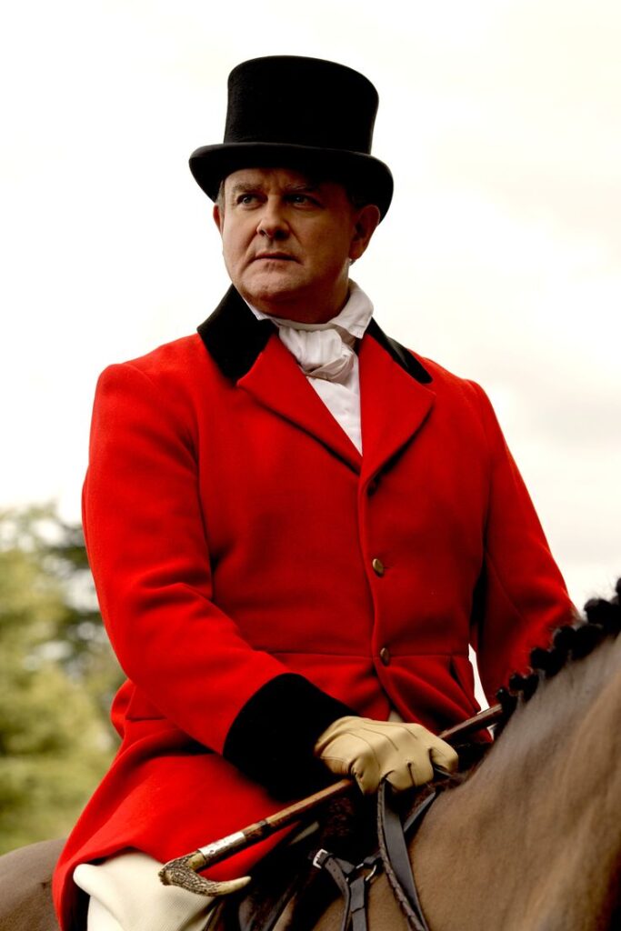 Lord Grantham on a horse in a red blazer light trousers and top hat